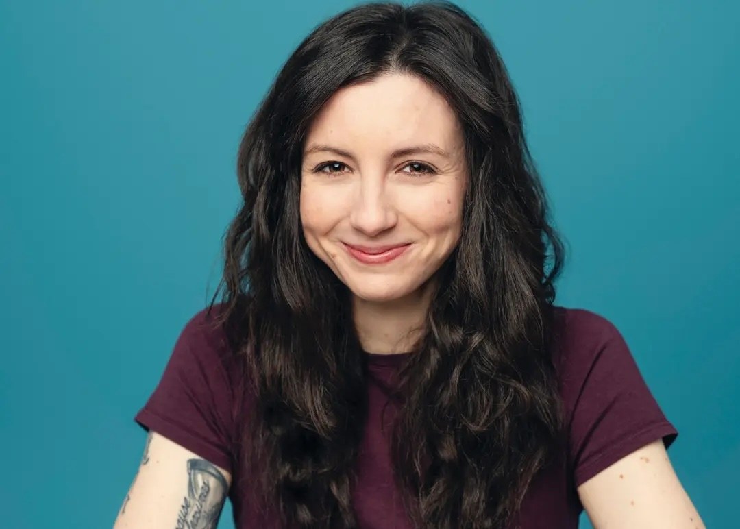 Cat McDonnell smiling, with long dark wavy hair, wearing a maroon t-shirt against a teal background